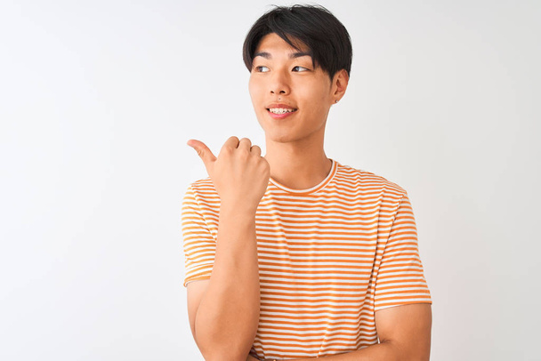 カジュアルなストライプのTシャツを着た若い中国人男性は、孤立した白い背景の上に立ち、幸せそうな顔で微笑み、親指を上げて横を向いている. - 写真・画像