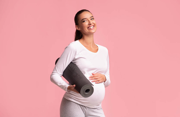 Terhes lány gazdaság jóga matrac álló felett rózsaszín háttér, Studio - Fotó, kép