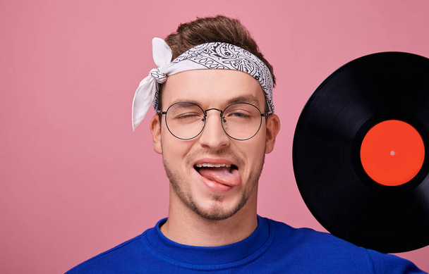 Glad hippie guy with protruding tongue and covered right eye in eyeglasses with black frame in white bandana holds at left hand one black vinyl record with a red label on the shoulder. Large frame. - Fotografie, Obrázek
