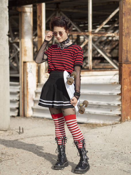 Alternativa ragazza su uno sfondo di costruzione abbandonato togliendo hipster occhiali rotondi e tiene bianco penny skate board
 - Foto, immagini