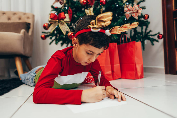 Boy lying on the floor under the Christmas tree writing wish list for Santa. Waiting for Christmas. Celebration. New Year. - Fotó, kép
