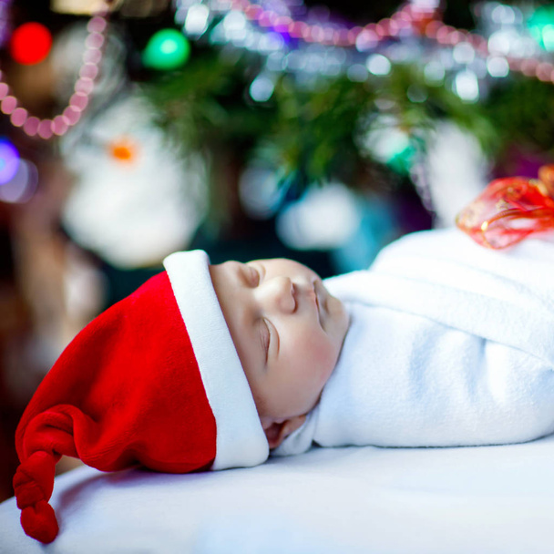One week old newborn baby in Santa hat wrapped in blanket near Christmas tree with colorful garland lights on background. Closeup of cute child, little baby sleeping. Family, Xmas, birth, new life. - Foto, afbeelding