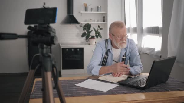 modern pensioner blogger Influencers works at a laptop computer and writes a vlog on mobile phone explaining bookkeeping to his followers - Footage, Video