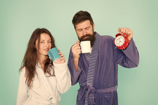 Man with beard and sleepy woman enjoy morning coffee or tea. Time to wake up and have nice day. Guy husband hold coffee and clock. Every morning begins with coffee. Couple in bathrobes with mugs - Foto, imagen