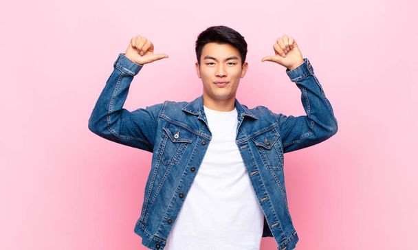 young chinese man feeling proud, arrogant and confident, looking satisfied and successful, pointing to self against flat color wall - Foto, Imagem