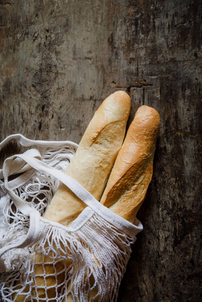 Fresh rosy baguettes in a string bag on a dark wooden retro background. Top view, copy space - Photo, Image