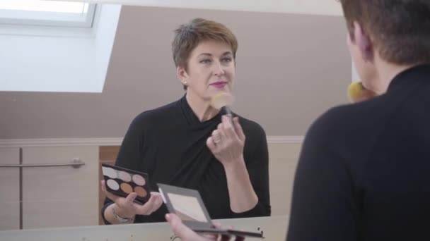 Close-up reflection in mirror of senior Caucasian woman looking at big mirror and applying face powder. Beautiful mature lady doing makeup. Fashion, beauty, face care, skin care. - Filmati, video