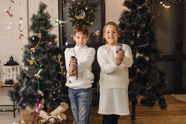 Δύο χαριτωμένα παιδιά στο φόντο των χριστουγεννιάτικων δέντρων ανοίγουν κράκερ και διασκεδάζουν. Αδελφός και αδελφή σε λευκό πλεκτό πουλόβερ γιορτάζουν την Πρωτοχρονιά και τα Χριστούγεννα σε ένα φωτεινό σπίτι - Φωτογραφία, εικόνα