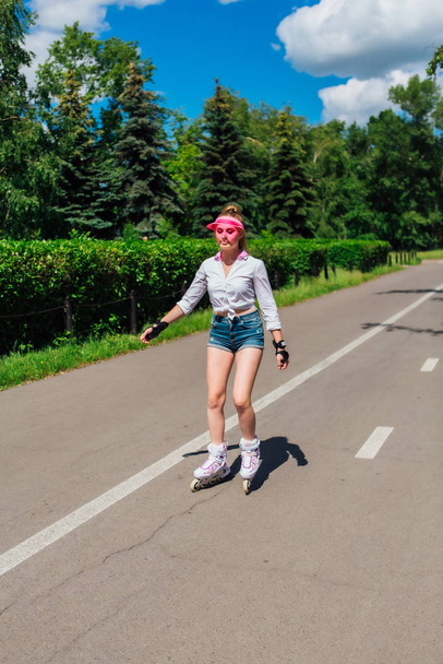 Portrait of an emotional girl in a pink cap visor and protective gloves for rollerblades and skateboarding riding on rollerblades on the road. - Photo, image