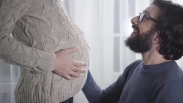 Side view of smiling Caucasian man in eyeglasses caressing his pregnant wifes belly and listening to babys heartbeat. Happy father waiting for his girlfriend to give birth. Pregnancy, gestation. - Кадри, відео