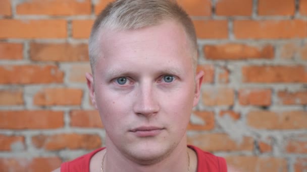 Portrait of handsome man with blonde hair looking into camera with serious and confident expression on his face outdoor. Close up of strong young sportsman standing on blurred brick wall background - Footage, Video