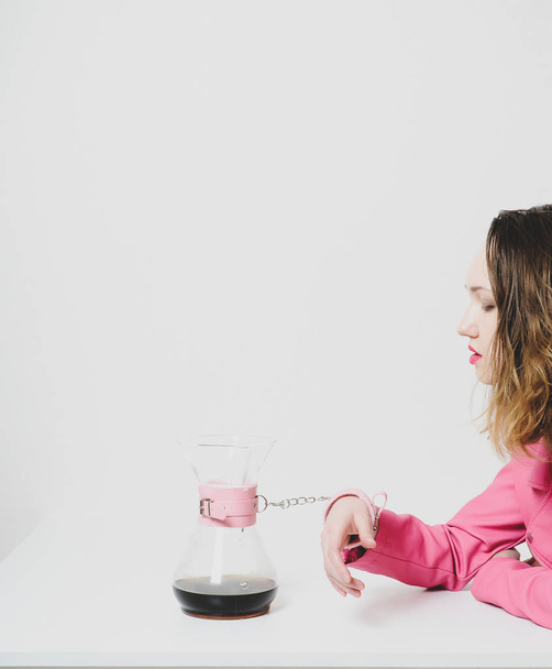 Ένα όμορφο κορίτσι με ροζ μπουφάν είναι δεμένο με ροζ χειροπέδες σε μια γυάλινη κανάτα καφέ. Μεταφορά πάθους, αλληγορία στοργής, εθισμός στην καφεΐνη. Αισθητική εικόνα - Φωτογραφία, εικόνα