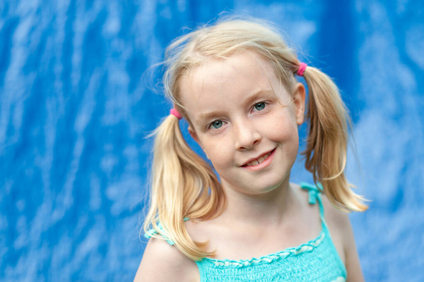Pretty young blonde girl with pigtails making eye contact on a blurry blue background - Photo, Image