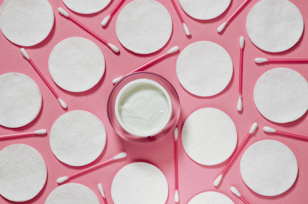Προϊόντα υγιεινής: λευκά στρογγυλά βαμβακερά μαξιλάρια και πλαστικά sticks για τον καθαρισμό των αυτιών βρίσκονται σε ροζ φόντο δίπλα στην κρέμα προσώπου - Φωτογραφία, εικόνα