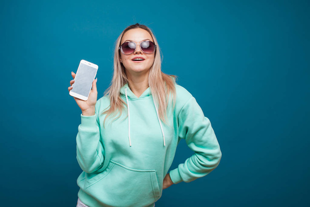 Stylish trendy hipster girl in the blue hoodie with the hood uses a smartphone. - Фото, изображение