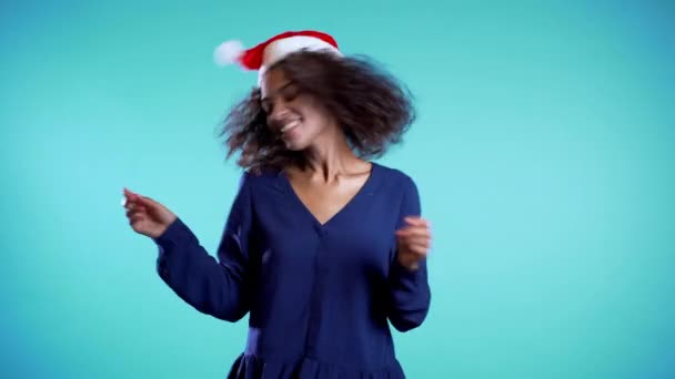 Attractive mixed race girl in Santa hat celebrating Christmas, New year on blue - Imágenes, Vídeo