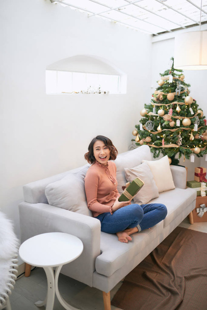 Beautiful asian woman in casual sweater holding gift near decorated christmas tree, greeting card concept - Photo, image