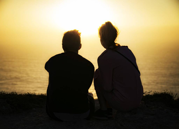 A silhouette of a young couple sitting on the beach enjoying the beautiful view of the sunset reflected in the sea - Fotó, kép