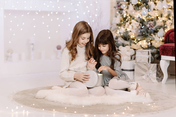 Счастливые две маленькие сестры открывают волшебный рождественский подарок у камина в уютной светлой гостиной накануне Рождества. Зимний вечер дома с семьей и детьми
. - Фото, изображение