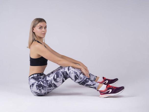 A sporty lady sitting with a straight posture with a straight look to forward. Sits on the floor in white cyclorama background. Sports top and leggings - Foto, Imagen
