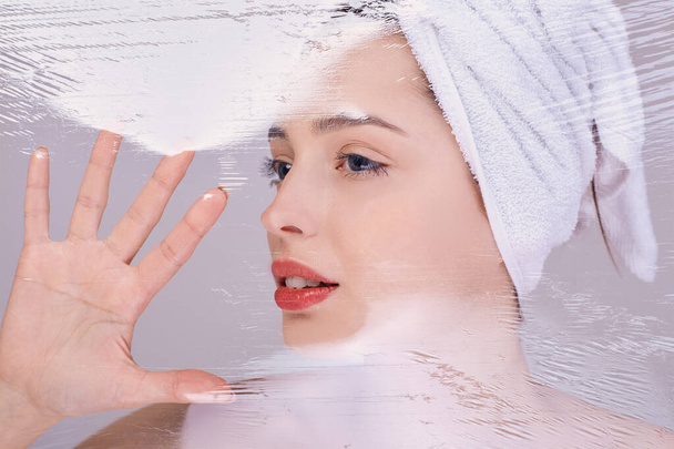 Young beautiful girl after a shower, with a towel on her head, tries to break through the cellophane. Silicone cosmetics. Natural cosmetic. The concept of health and beauty. Organic cosmetics. - Foto, afbeelding