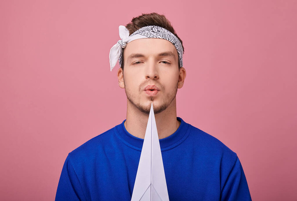 Close up. Calm stylish guy in a white with patterns bandana in dark blue sweater is standing with white paper airplane in his hand, that shows to the side. Boy blowing on an airplane. Copy space. - Фото, изображение
