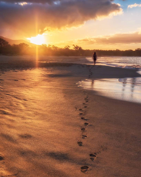 A vertical shot of a female walking through the ocean shore leaving traces on the wet sand at sunset - Foto, imagen
