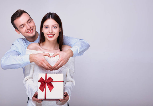 A cheerful young man in blue shirt is standing behind cute attractive smiling girl in snow-white sweater with gift in her hands for Valentine's Day. Guy shows heart sign with his own hands. Copy space - Photo, Image