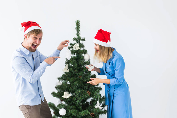 Праздники и праздничная концепция - Молодая семейная пара в шляпах Санта-Клауса, украшающая елку
 - Фото, изображение