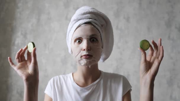 A woman in a face mask is having fun with round cucumbers, looking at the camera and fooling around. Natural Regenerating Skin Care - Materiał filmowy, wideo