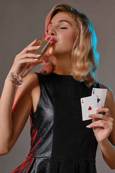 Blonde girl in black dress holding two playing cards and drinking champagne, posing against gray background. Gambling, poker, casino. Close-up. - Photo, Image