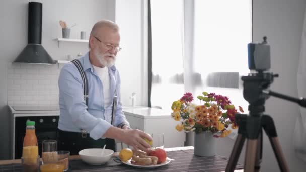 řádná výživa, usmívající se milý muž blogger říká svému předplatiteli recept na zdravé jídlo tím, že natáčí video na svém mobilním telefonu pro vlog, zatímco stojí ve své kuchyni - Záběry, video
