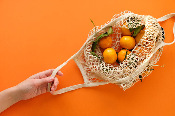 A human hand holds a string bag with tangerines on bold orange color background. - Фото, зображення