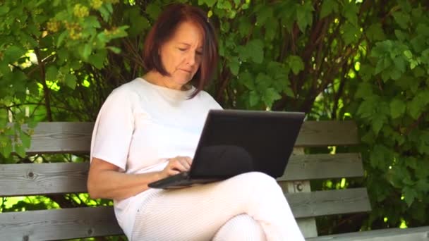 Střední dospělá žena pracující venku na notebooku, usmívající se babička středního věku pracující vzdáleně na počítači. - Záběry, video