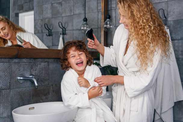 Charming woman dressed in white bathrobe combs her sons curly hair while he sitting on washbasin and laughing. - Foto, Imagem