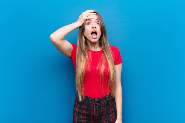 young pretty woman panicking over a forgotten deadline, feeling stressed, having to cover up a mess or mistake against blue wall - Fotoğraf, Görsel