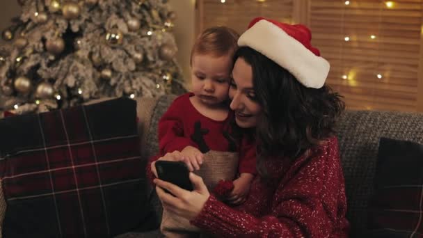 Smiling Happy Mother Wearing Santas Hat with her Cute Baby Son Sitting on the Sofa in Living Room near Christmas Tree Using Looking at Smartphone Concept of Holidays and New Year. - Záběry, video