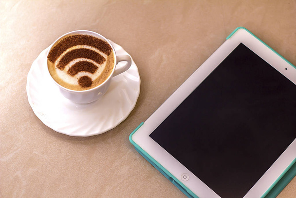 A cup of cappuccino coffee with latte art on milk foam with a wi-fi symbol and tablet pc  - Fotografie, Obrázek