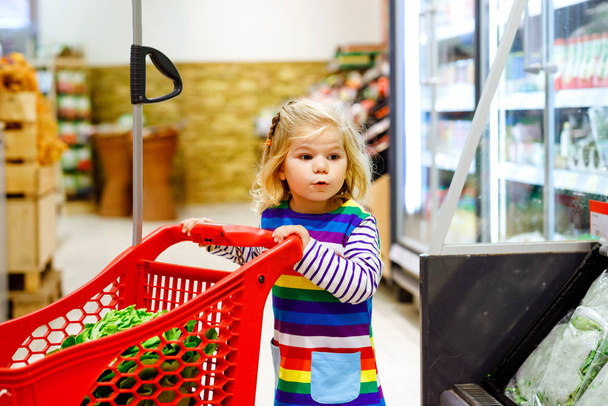 Cute todler girl pushing shopping cart in supermarket. Little child buying fruits. Kid grocery shopping. Adorable baby kid with trolley choosing fresh vegetables in local store. - Photo, Image