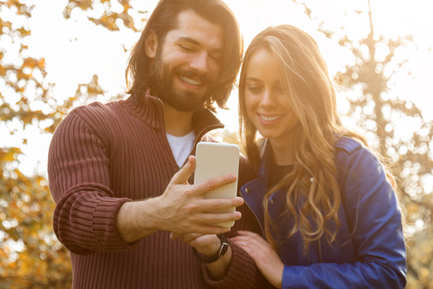 Молодая пара использует мобильный телефон в осеннем цветном парке
. - Фото, изображение