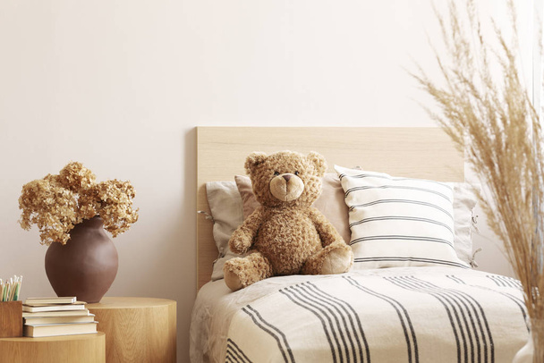 Flowers in brown vase on wooden nightstand table next to single bed with stripped bedding with teddy bear - Photo, Image