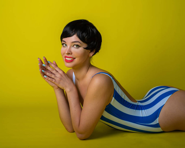 Attractive brunette lies on her stomach in a striped bikini. Short-haired young woman in bodysuit on a yellow background in the studio. - Φωτογραφία, εικόνα