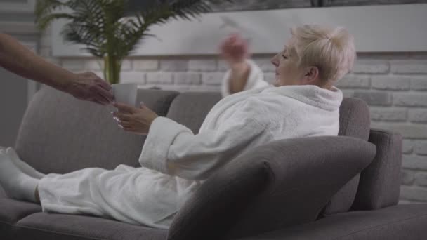 Side view of mature ill Caucasian woman lying on couch in white bathrobe. Her adorable baldheaded husband coming up with hot drink and pills. Aging, healthcare, eternal love, care. - Záběry, video