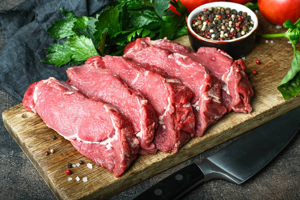 frisches rohes Rindfleisch, geschnitten in Steaks auf einem Holzbrett mit Gemüse, Kräutern und Gewürzen auf einem Holzbrett auf dunklem Hintergrund. - Foto, Bild