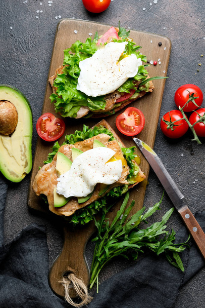 leckeres Sandwich mit Avocado und pochiertem Ei, Kirschtomaten und Rucola auf dunklem Hintergrund. Ansicht von oben.  - Foto, Bild
