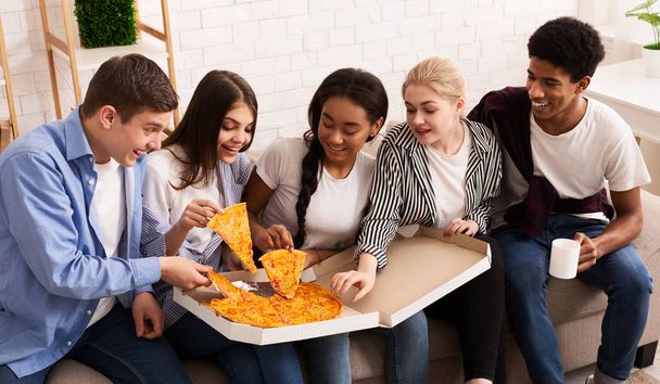 Nastoletni współlokatorzy jedzą gorącą pizzę, biorą kawałki z pudełka. - Zdjęcie, obraz