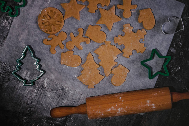 vykrojená perníková sušenka ve formě vánočního stromku, hvězdy, mužíčka, srdíčka ze syrového těsta na pergamenu pečící papír na tmavém pozadí. Horní pohled. Uložit místo - Fotografie, Obrázek