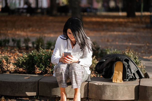 Attractive girl resting on nature at weekend. Girl in white shirt sitting in park. Caucasian brunette young woman sending message on smartphone outside alone.Technologies and gadgets.Horizontal - Foto, immagini