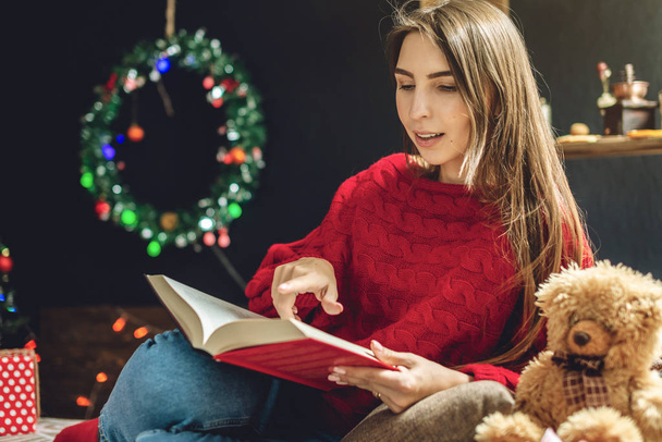 Kobieta w czerwonym swetrze czytająca wieczorem książkę w ciepłej świątecznej atmosferze. Przytulny nastrój nowego roku - Zdjęcie, obraz