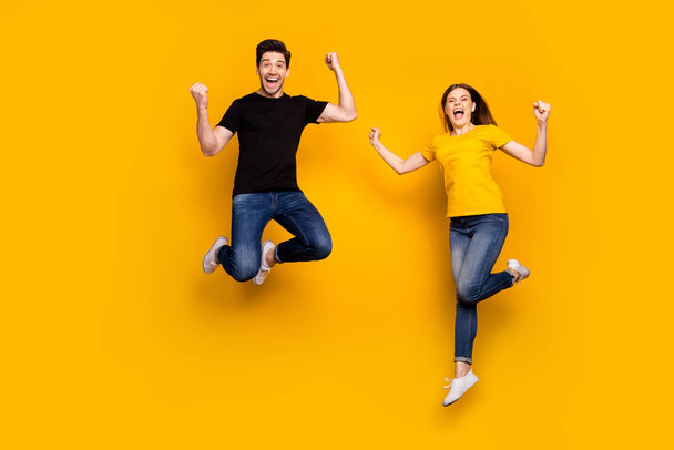 Full body photo of crazy guy lady couple jumping high celebrating first win place competition triumphing wear casual jeans black t-shirts isolated yellow color background - Zdjęcie, obraz